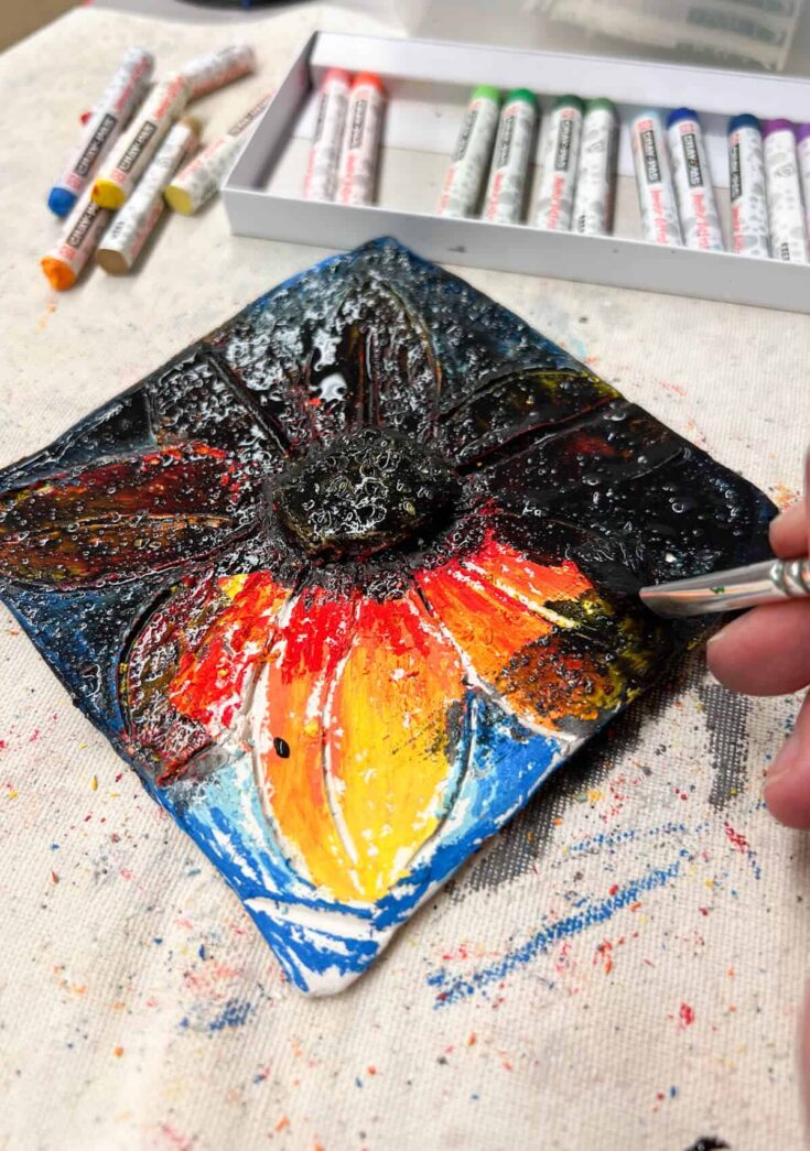 black paint being brushed on clay tile covered with oil pastels.