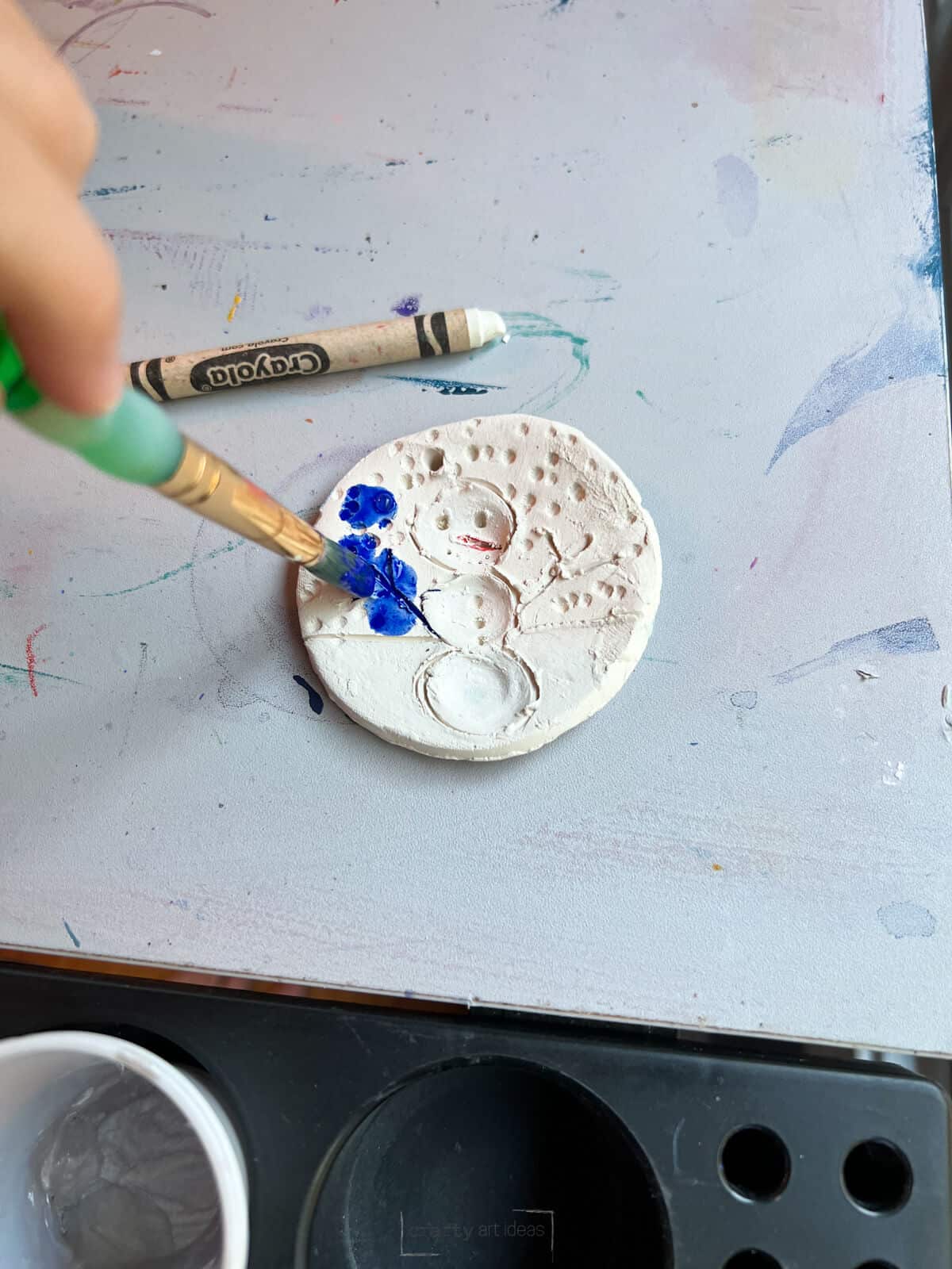 kid painting blue watered down tempera paint on clay snowman ornament.