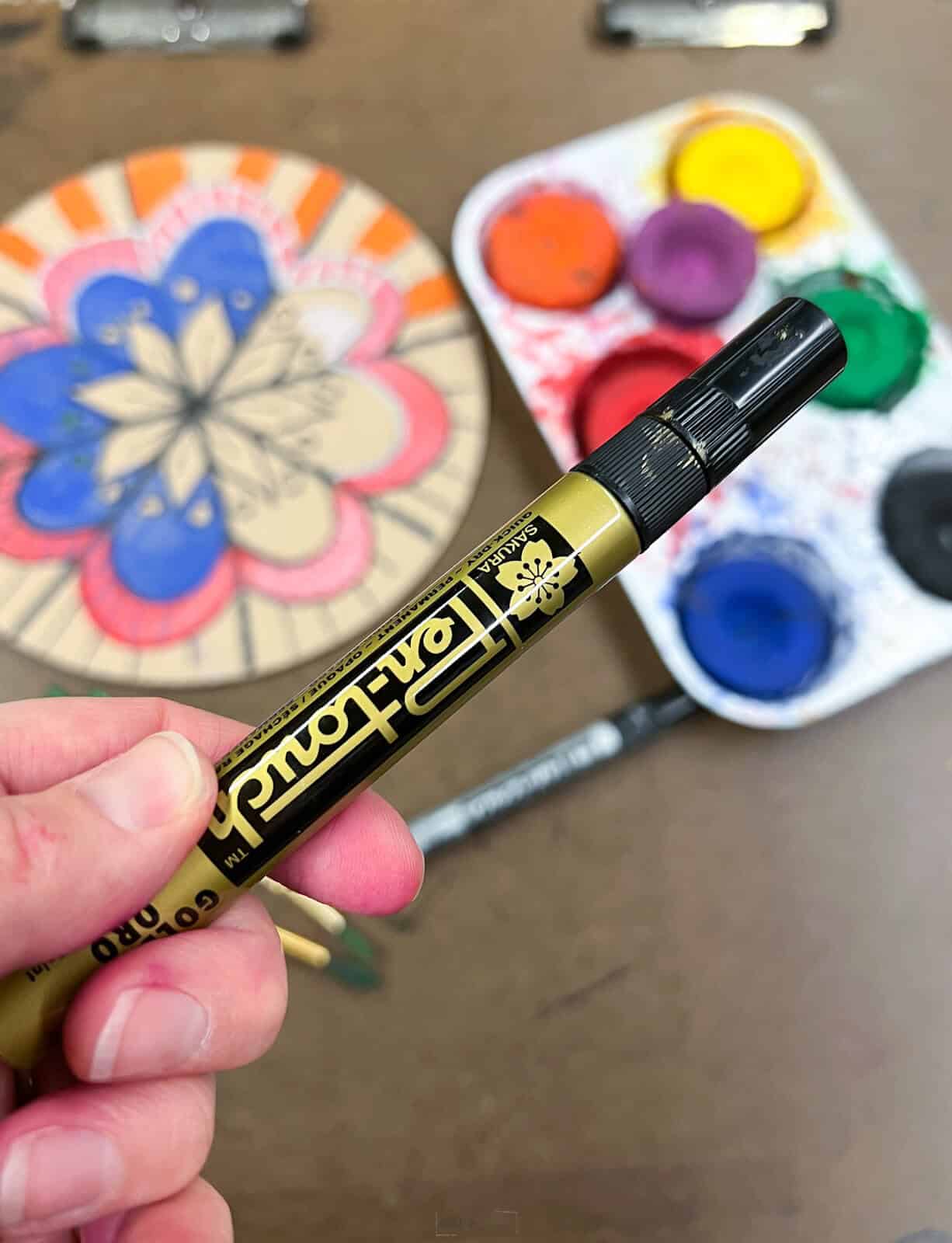 hand holding gold paint marker with mandala design and paints in the background.