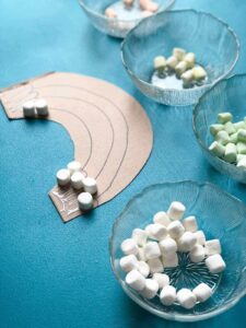 cardboard rainbow with glue and marshmallows with bowls of colored marshmallows on teal table.