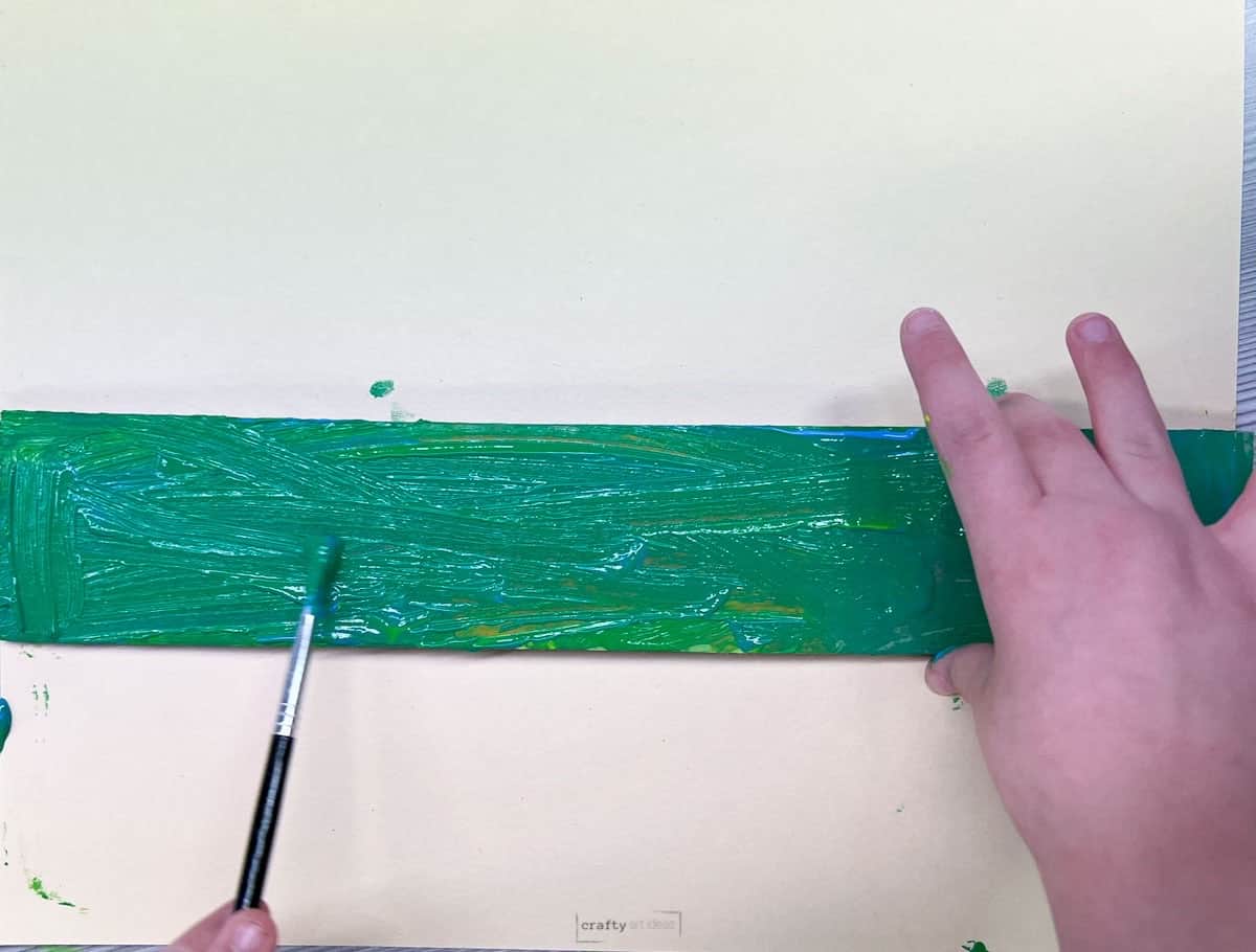child's hand holding paint brush painting a cardboard strip green.