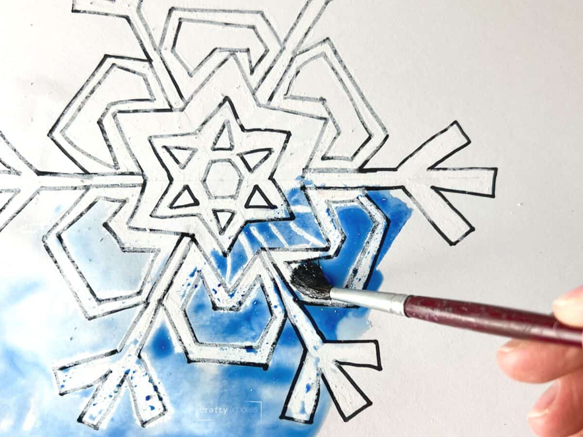 hand holding paintbrush painting blue watercolor paint on top of white oil pastel snowflake.