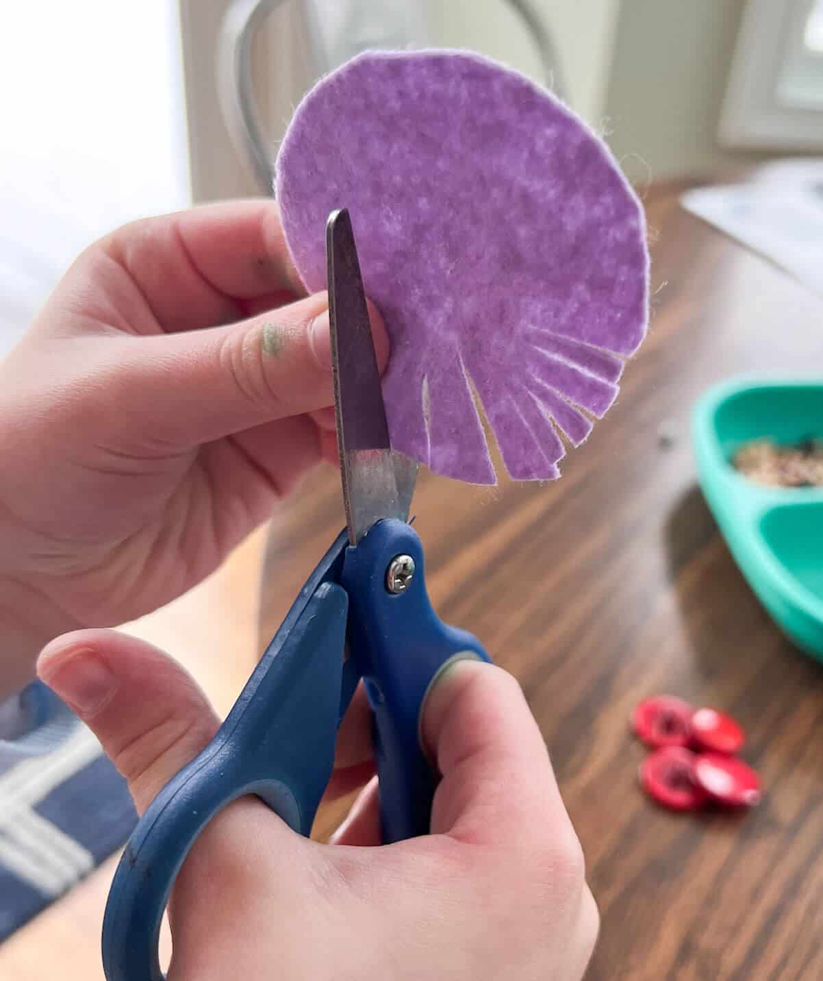 child's hand using blue scissors to cut fringe in purple felt circle.