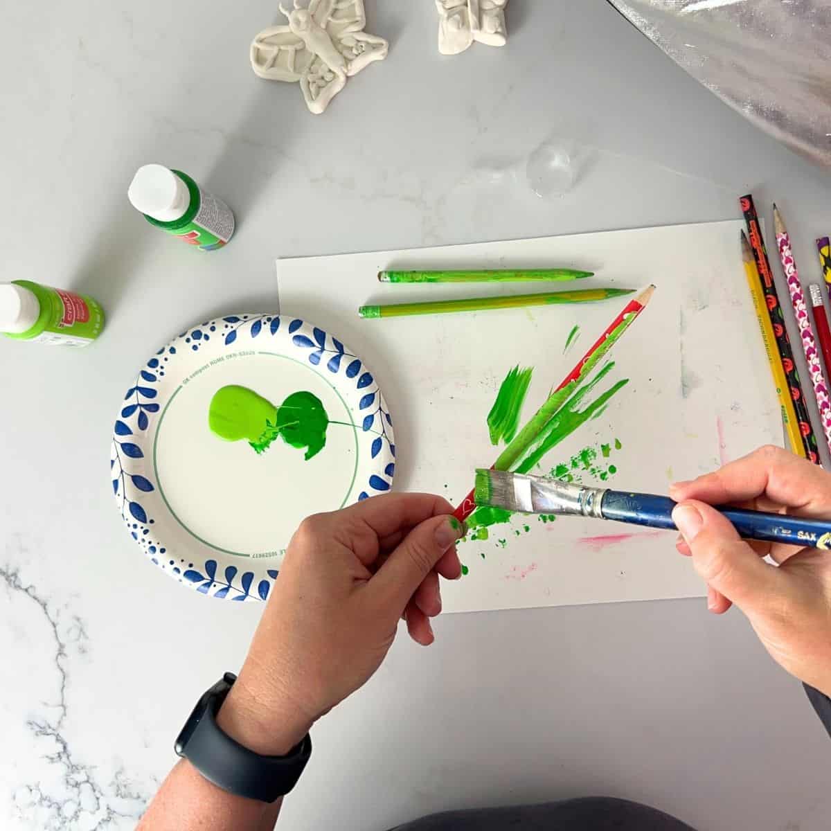 hand holding paint brush adding green paint to a pencil.