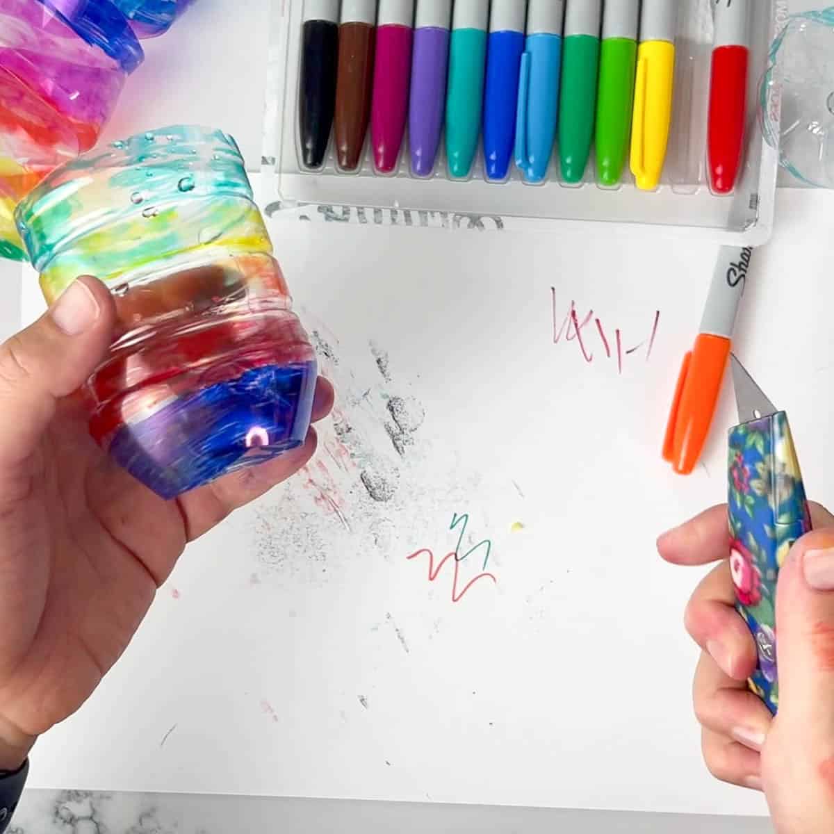 hand holding colorful bottle and the other hand holding utility knife.