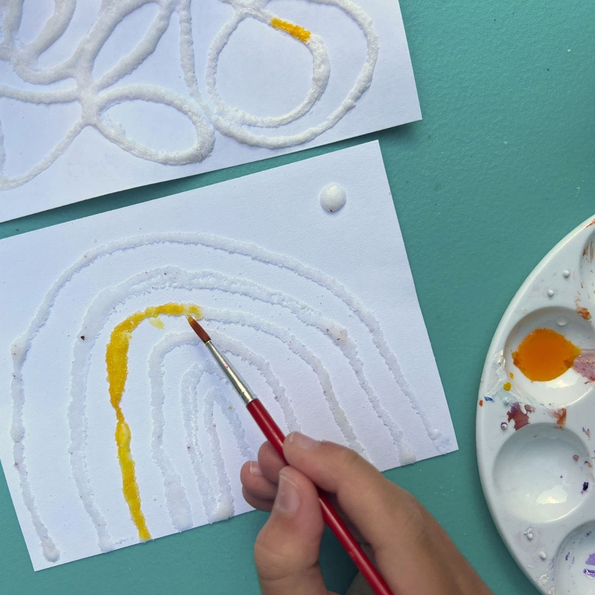child's hand using liquid watercolor paint to add yellow to rainbow.
