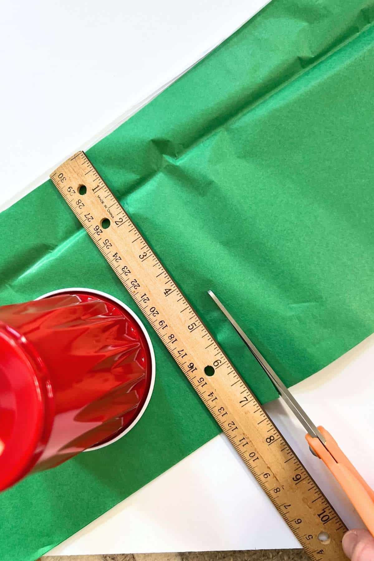 scissors cutting green tissue paper larger than a red cup.
