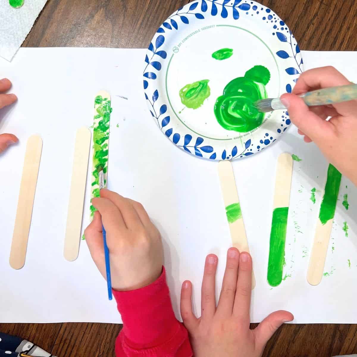 hand using paint brush to add green paint to popsicle craft sticks.