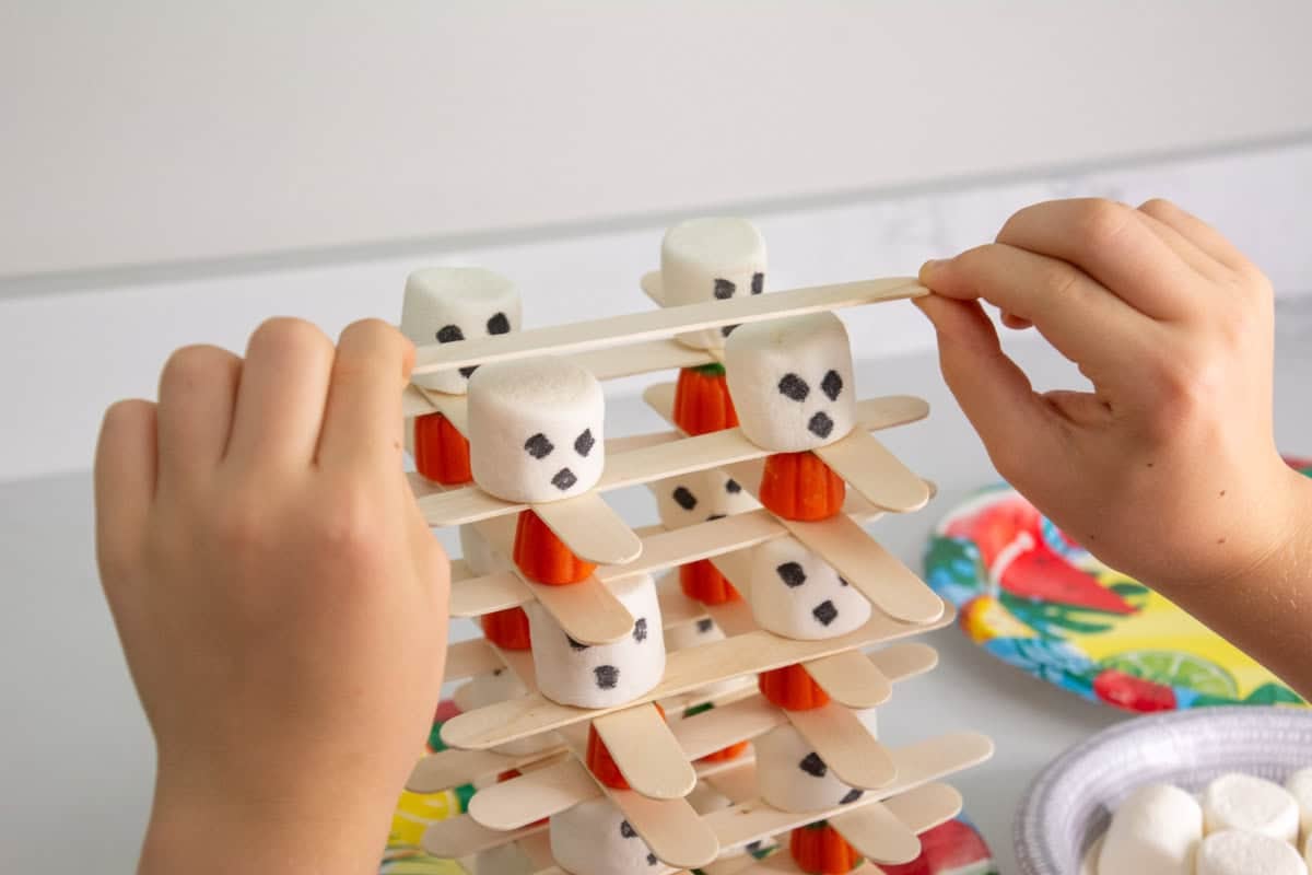child's hand stacking popsicle sticks on top of ghost marshmallows.