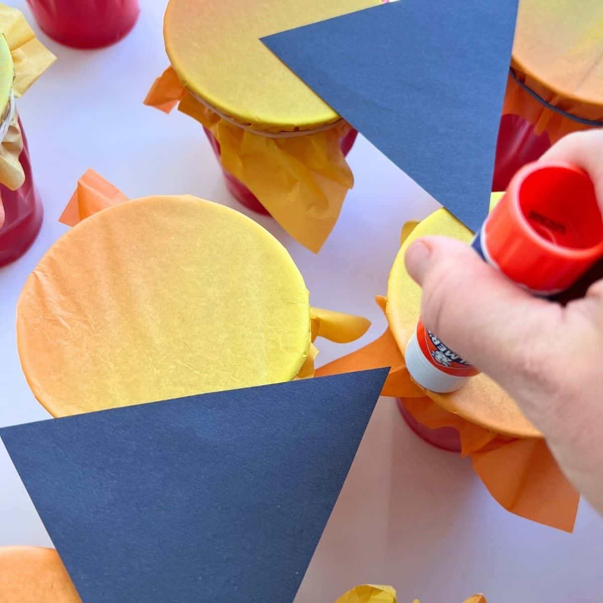 hand using glue stick to add black construction paper to orange tissue paper cups.