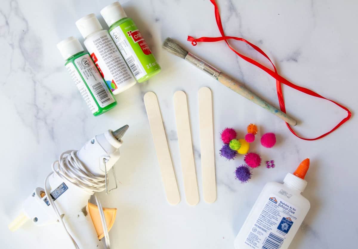 glue gun, regular glue, green and white paint bottles, 3 craft sticks and pom poms on a table.