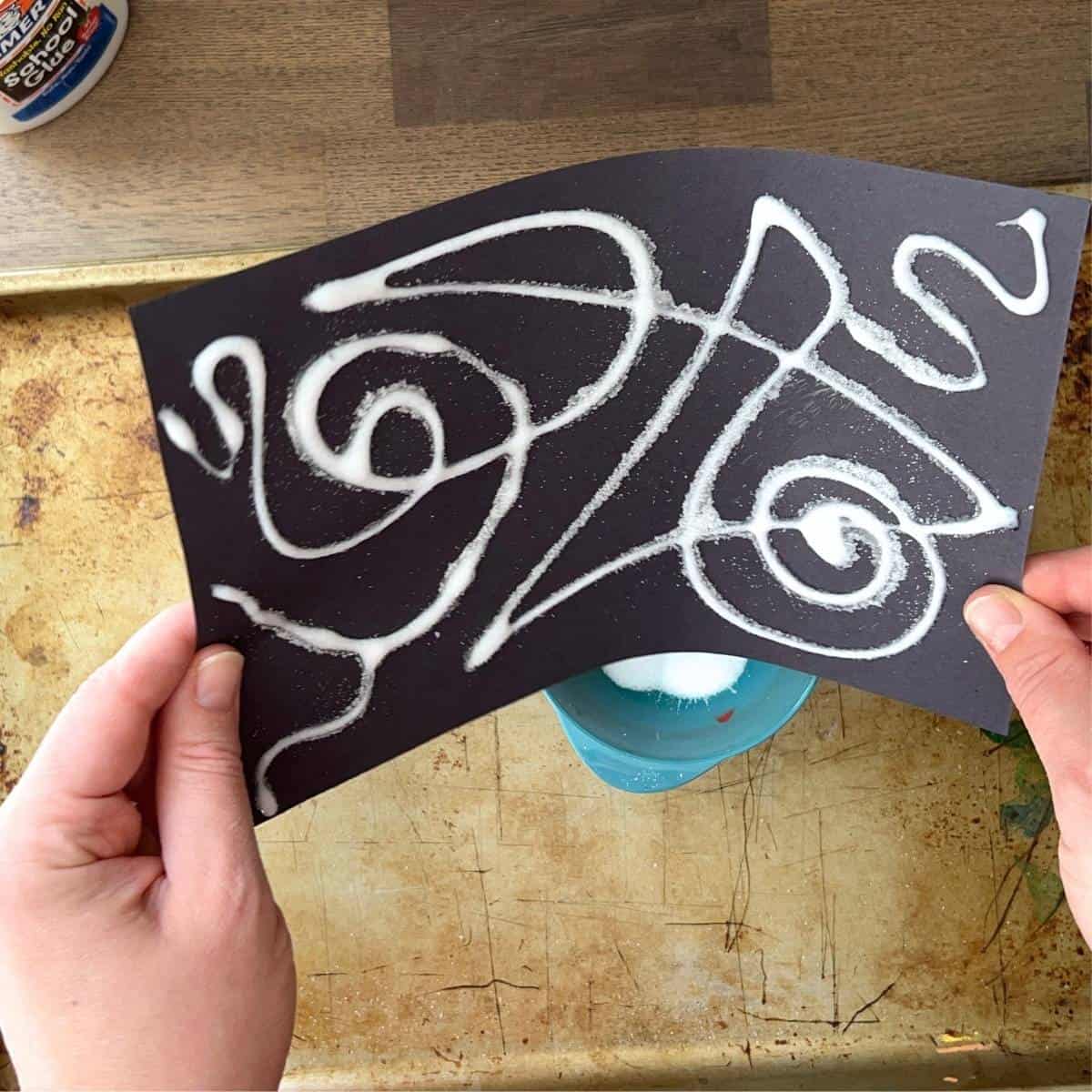 hand shaking black paper with glue and salt lines one it.