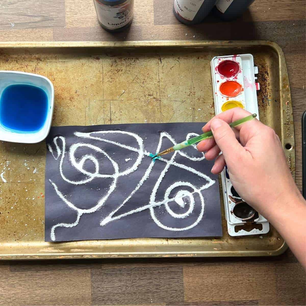 hand using paint brush to add blue watercolor paint to lines on black paper.