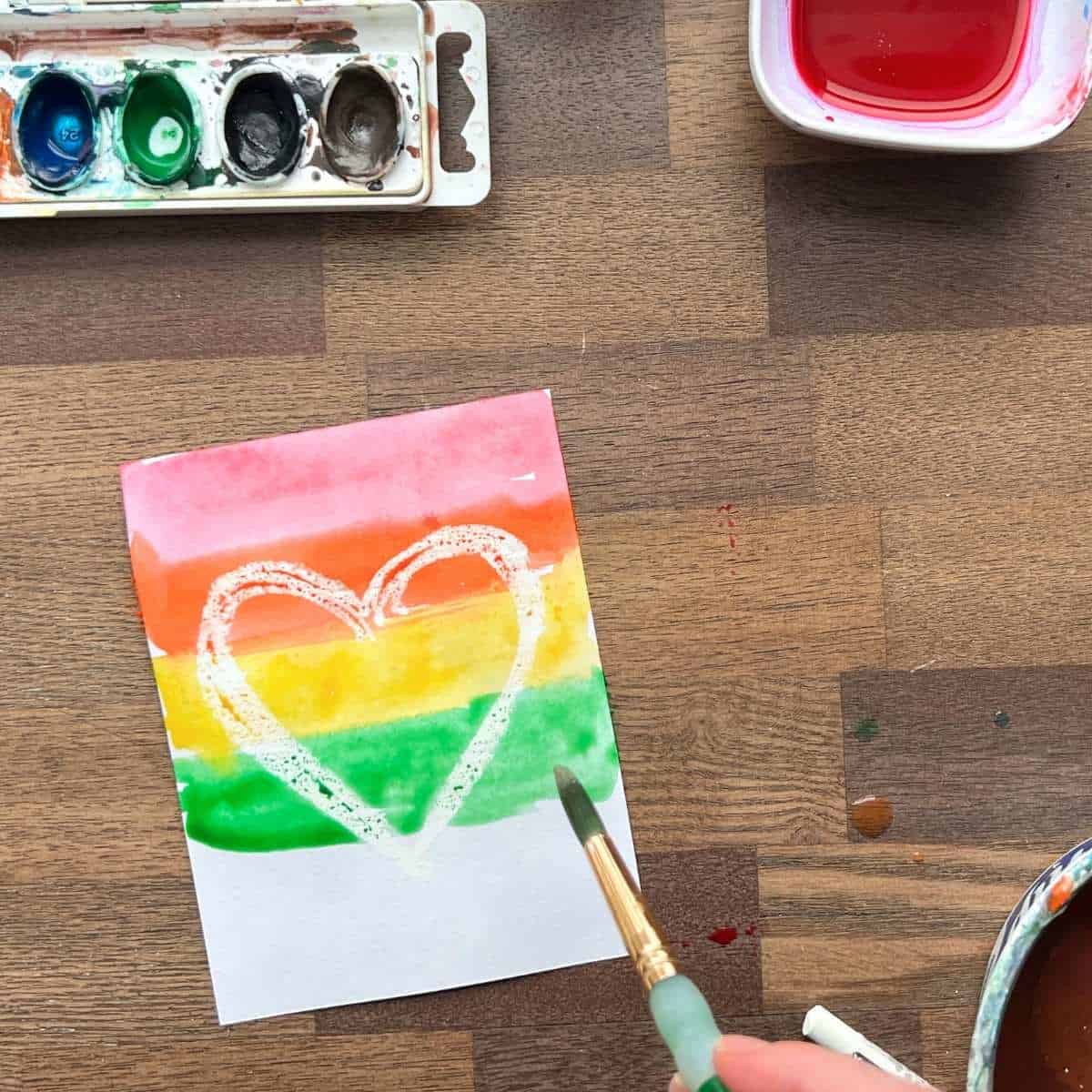 hand adding green watercolor paint to rainbow heart painting.