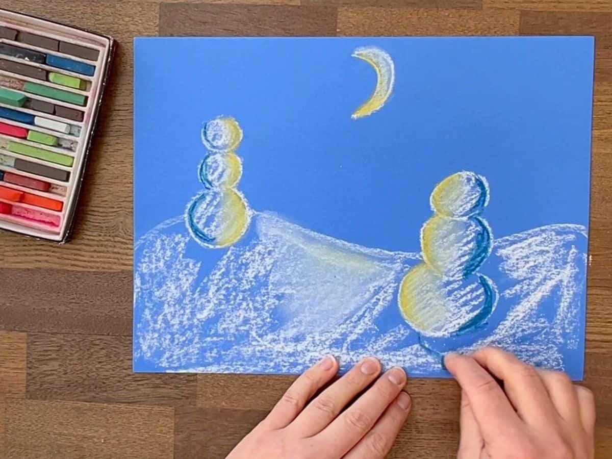 hand using darker blue pastels on snow drawing.