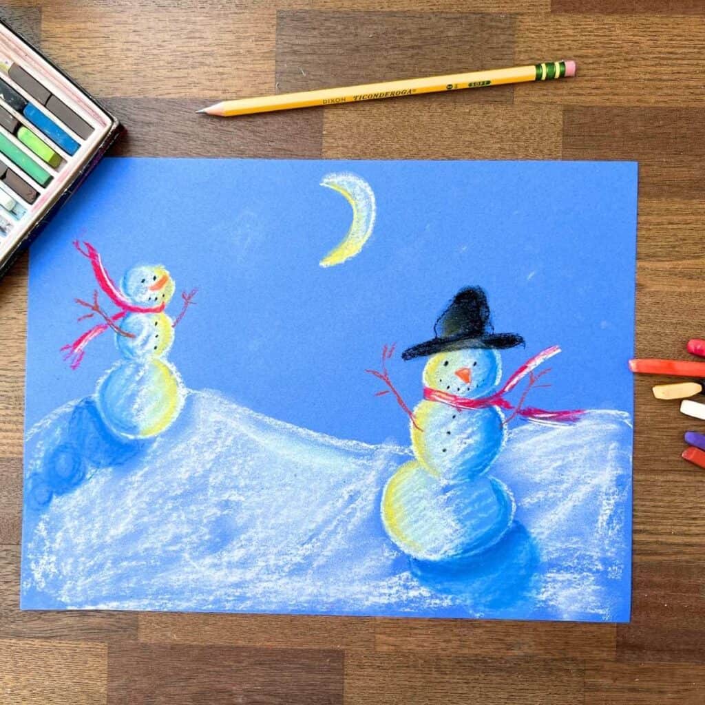 snowmen at night chalk drawing on blue paper sitting on a table.