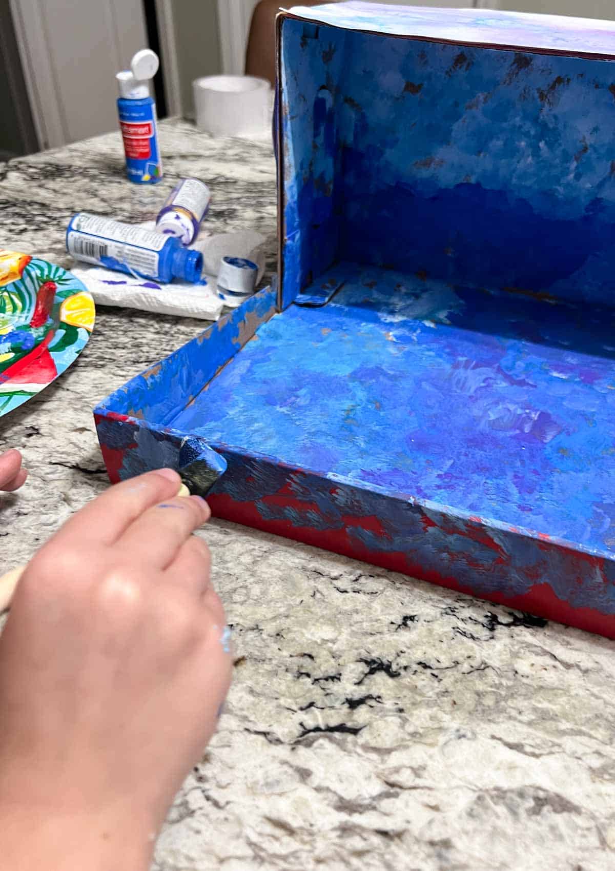hand using small brush to add blue paint to shoebox diorama.