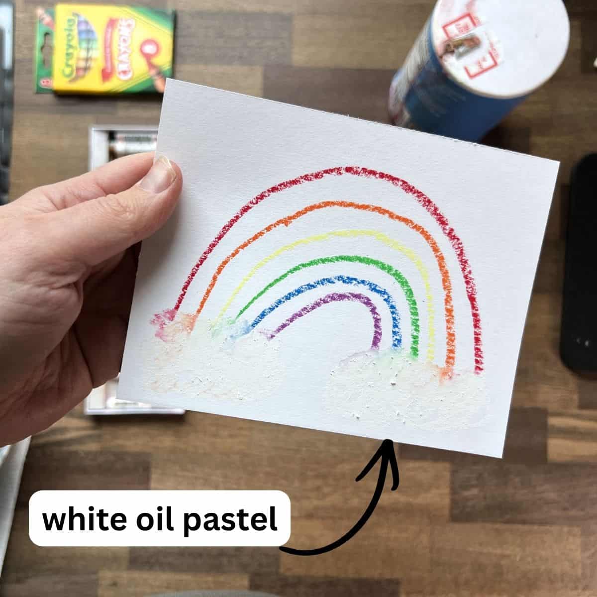 hand holding rainbow drawing with white oil pastel clouds.