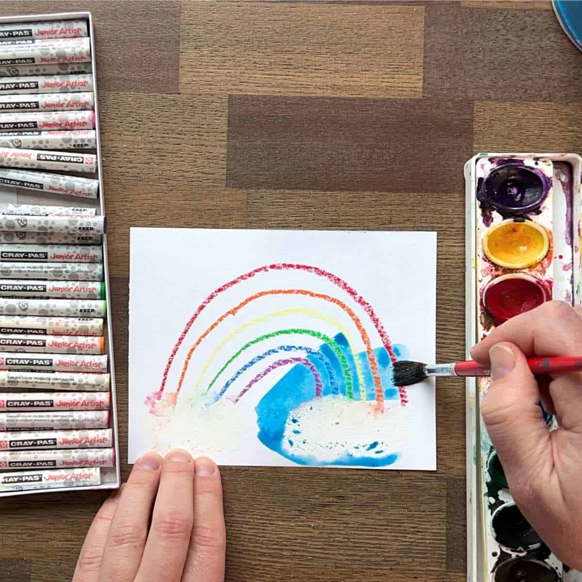 blue watercolor paint being adding by hands over a rainbow drawing.