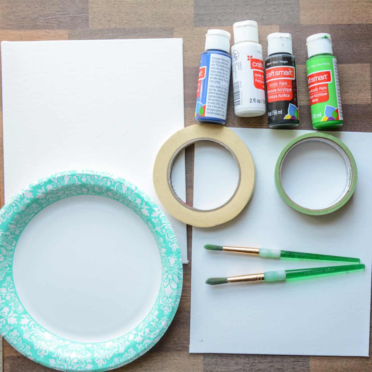 canvas boards, paper plate, craft paint, tape and paintbrushes on a table.