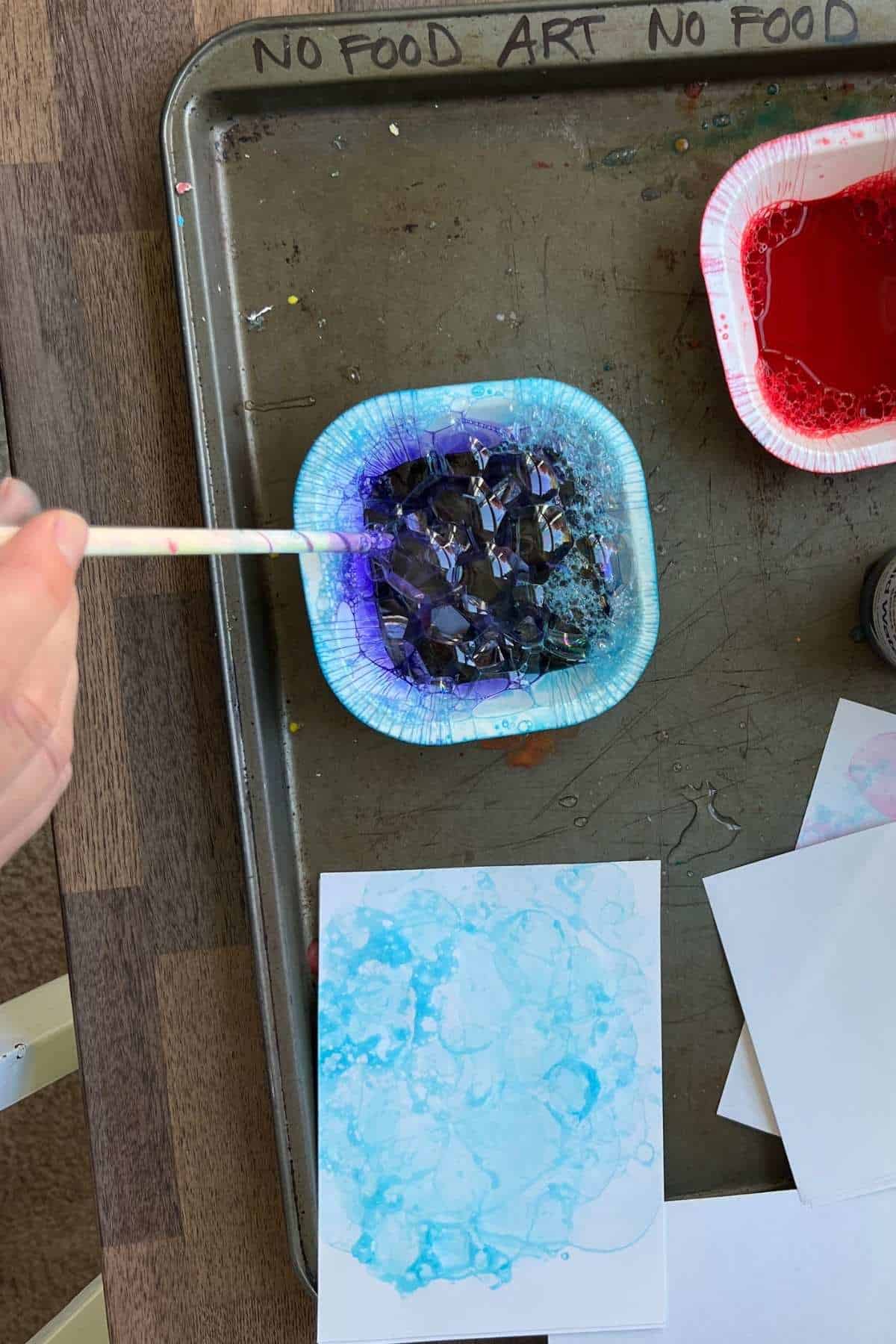 hand holding straw to blow bubbles in blue water paint.