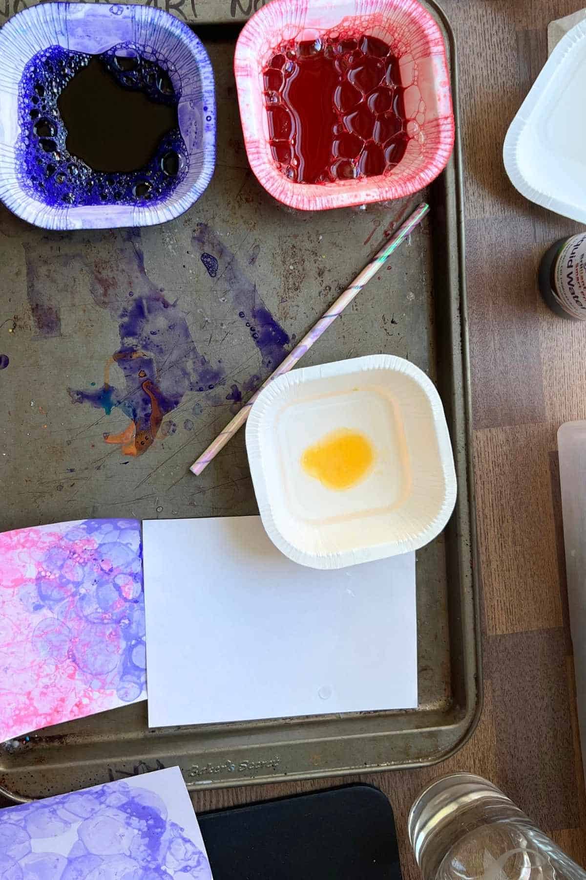 dish soap in shallow container with water and purple and red paint in a container.