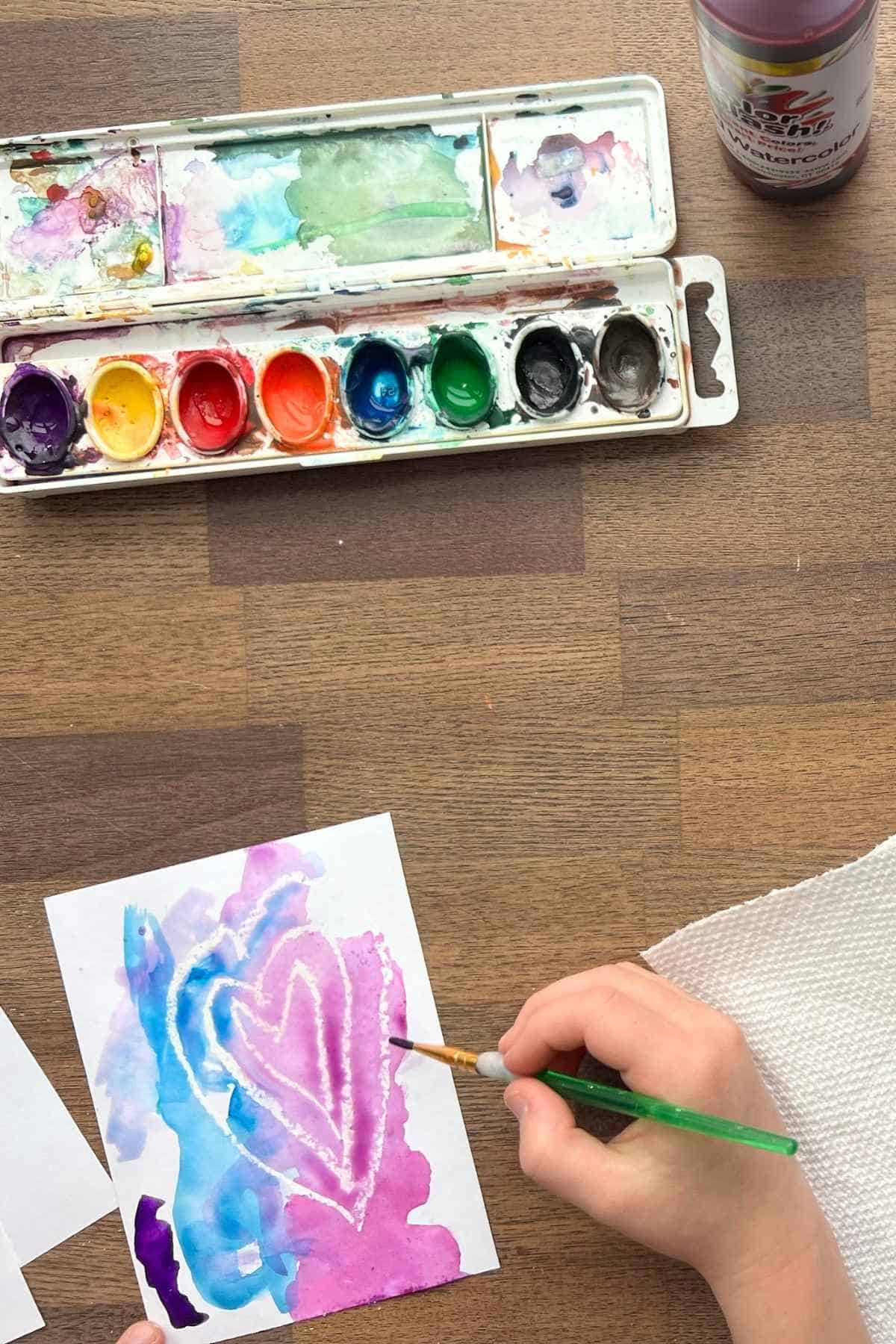 child's hand painting watercolor paint over white heart drawing.