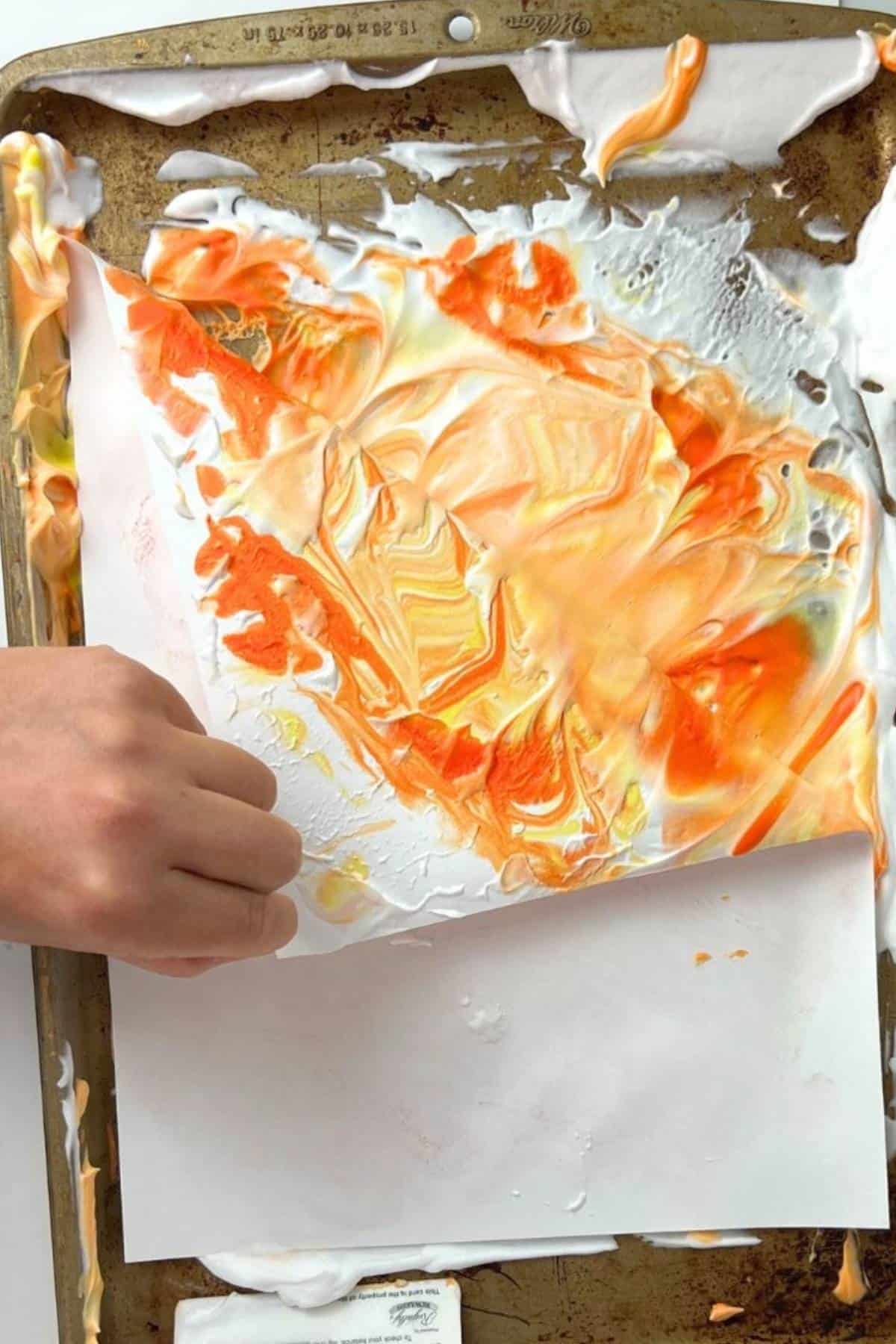 hand pulling paper off of colorful shaving cream on a tray.