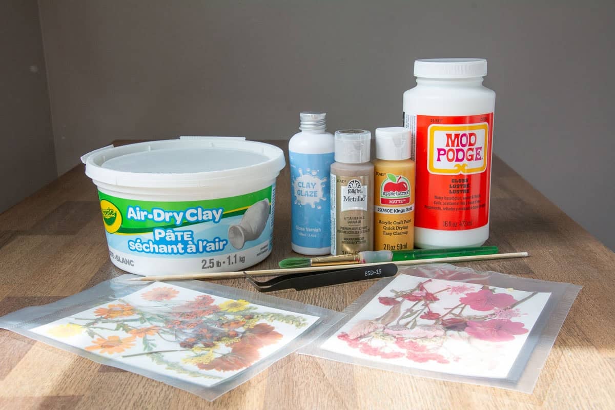 air dry clay, paints, clear coats and pressed flowers on paper on a table.