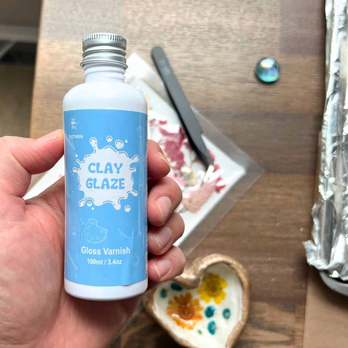 hand holding clear bottle of clear glaze with clay flower project on the table.